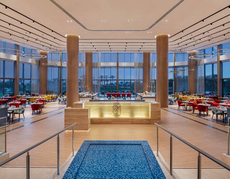 Stylish dining area at the JA Gallery with modern decor, red and yellow chairs, circular lights, and a prominent wooden panel centrepiece.