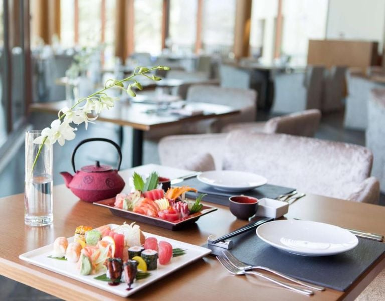 Table set with sushi and sashimi at JA Gallery, including a red teapot and white orchid flowers, in a modern dining room with elegant furnishings.