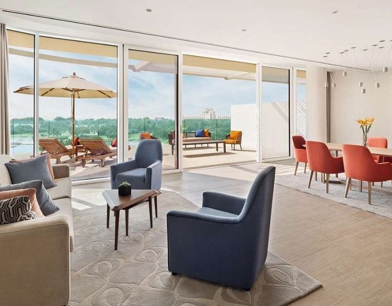 Modern living room with large glass doors, leading to a patio with loungers and a landscape view, featuring plush sofas, armchairs, and a dining table with red chairs.