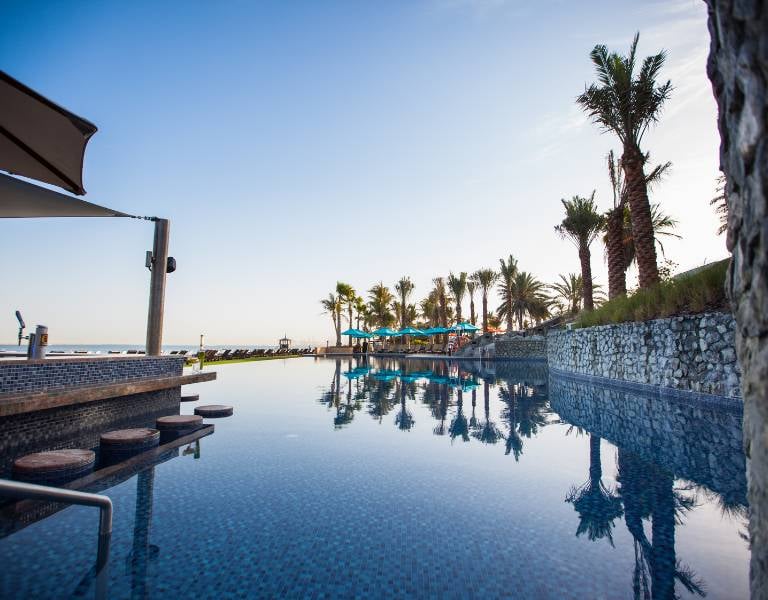 Infinity pool at JA Gallery Resort with ocean view, palm trees, and sun loungers.