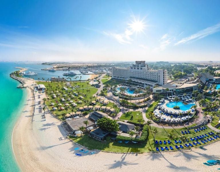 Aerial view of JA The Resort with pools, gardens, and a beachfront along turquoise waters under a sunny sky.