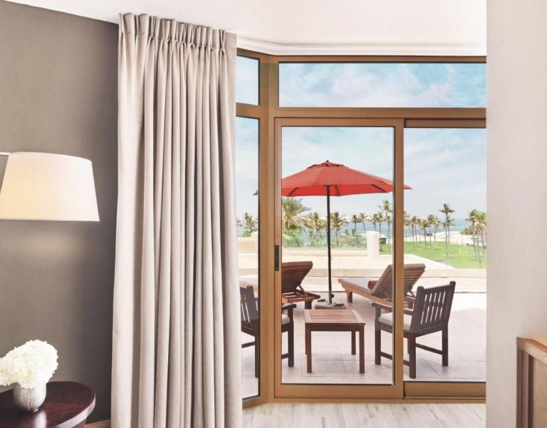 Hotel room at JA Resort with a large window opening to a patio with red umbrella and sea view.