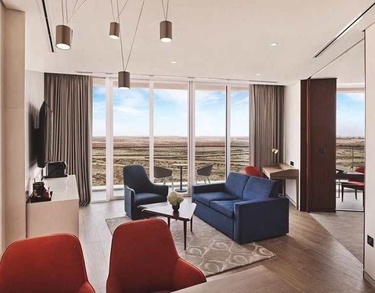 Modern hotel room with blue sofa, red chairs, large TV, and floor-to-ceiling windows overlooking a desert landscape.