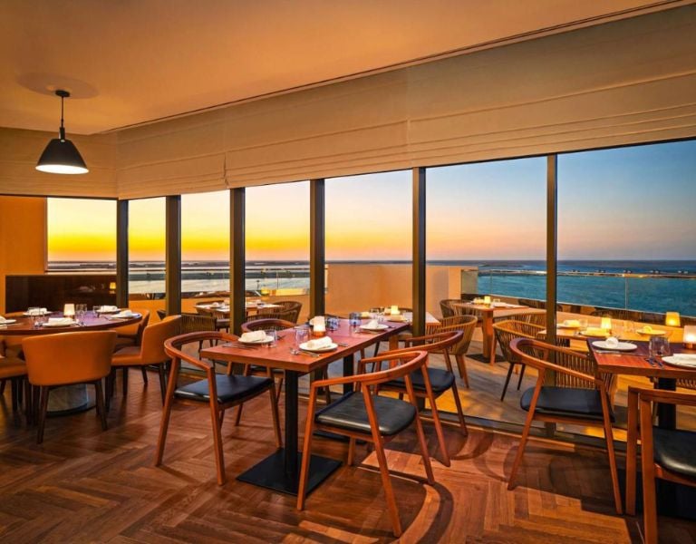 Restaurant interior with tables and chairs, large windows displaying a scenic ocean sunset view, creating a warm and inviting dining atmosphere.