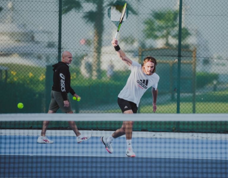 Male tennis player in a white Adidas outfit executing a forehand swing on an outdoor court with a coach in the background.