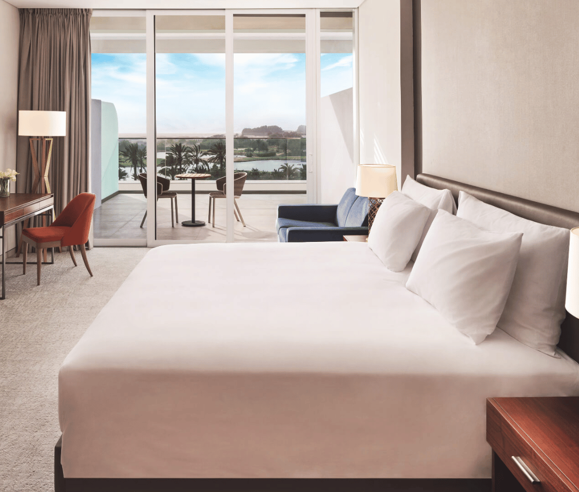 Deluxe room at JA Lakeview Hotel with a double bed, stylish furnishings, and a balcony view of palm trees and water.