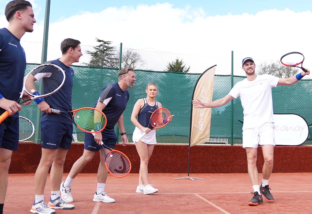 Jamie Murray greeting a group of tennis players on a clay court, all holding racquets and standing beside a promotional banner.