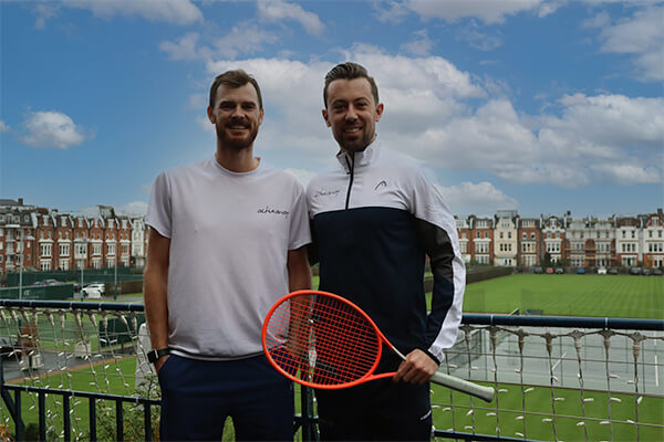 Jamie Murray with an Active Away representative holding a tennis racket on a balcony overlooking a tennis court and houses.
