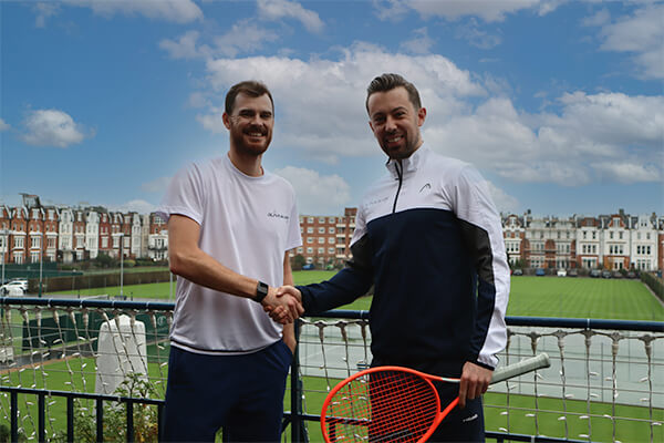 Jamie Murray and Josh Thompson shaking hands on a tennis court with a grass field and British houses in the background.