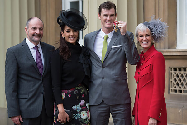 Jamie Murray OBE holds his OBE medal and stands with family members, all dressed elegantly in front of a grand building.