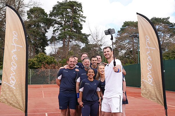 Jamie Murray with the Active Away team taking a selfie on a clay tennis court, framed by two Active Away flags.