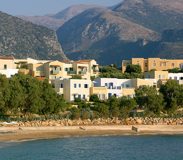 View of Kalimera Kriti Resort with pastel-hued buildings, lush greenery, and mountains, by the sea.