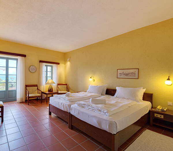 Hotel room at Kalimera Kriti with two double beds, yellow walls, wooden furniture, tiled floor, and natural light from the window.