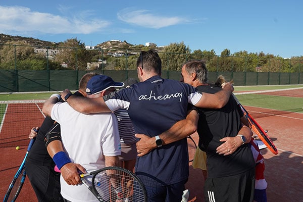 Active Away team huddle on Ikos Aria tennis court, with tennis rackets, under a clear blue sky.