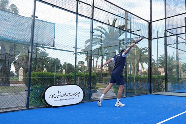 Active Away coach executing a backhand on a blue padel court with palm trees in the background.