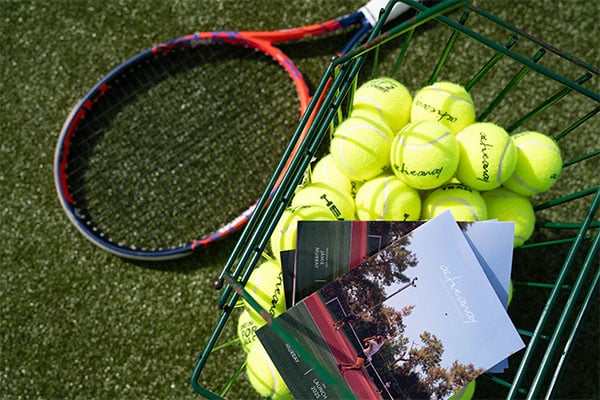 Tennis racket and basket of Active Away branded tennis balls with brochures on a grass court.