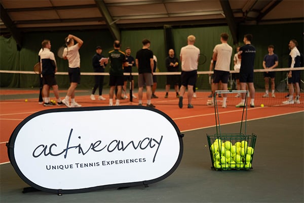 Indoor tennis court featuring a banner for 'Active Away' promoting unique tennis experiences and a basket of tennis balls, with participants in the background.