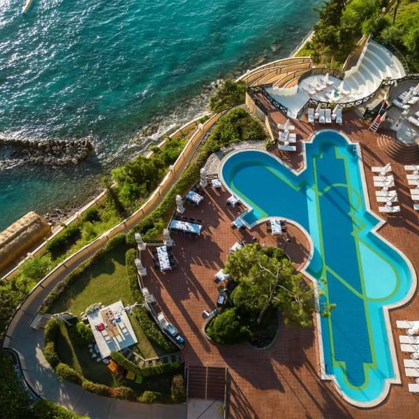 Aerial view of Liberty Lykia Resort's pool area, with a unique pool shape, surrounded by green landscaping and adjacent to the turquoise sea.