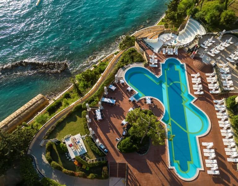 Aerial view of Liberty Lykia Resort's pool area, with a unique pool shape, surrounded by green landscaping and adjacent to the turquoise sea.