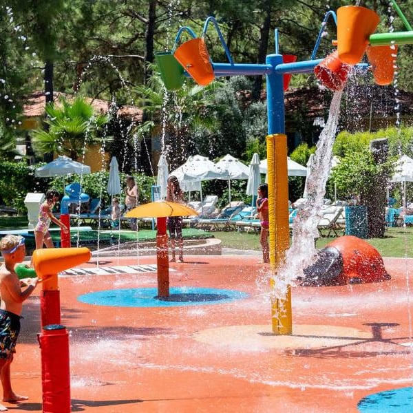Children playing in the colourful splash park at Liberty Lykia Family Resort, with water jets and buckets against a backdrop of greenery.