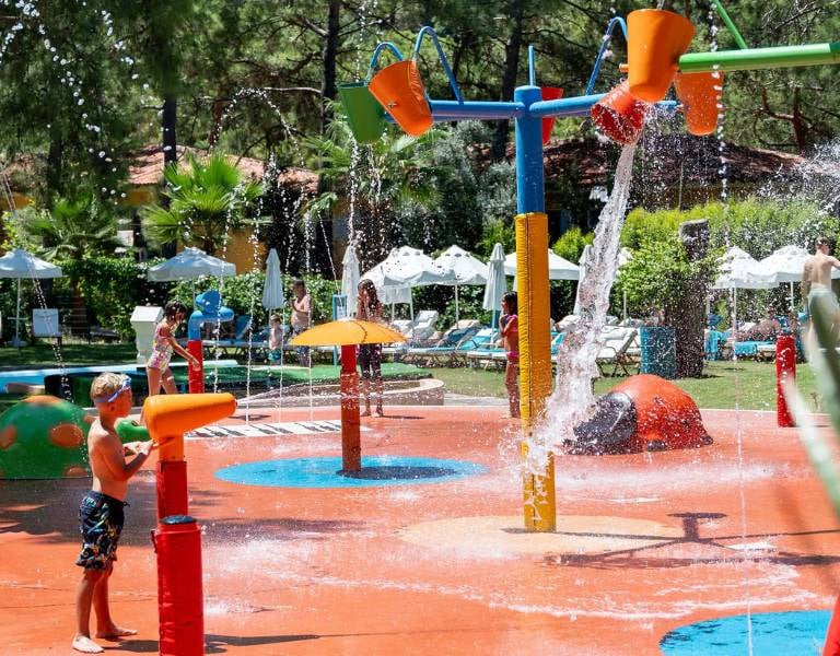 Children playing in the colourful splash park at Liberty Lykia Family Resort, with water jets and buckets against a backdrop of greenery.