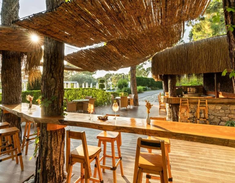 Outdoor bar at Liberty Lykia with wooden stools and bar, surrounded by greenery with woven shaded areas.