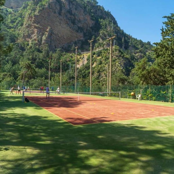 Tennis court at Liberty Lykia Resort with players, set against a backdrop of green mountains and trees, under a clear blue sky.