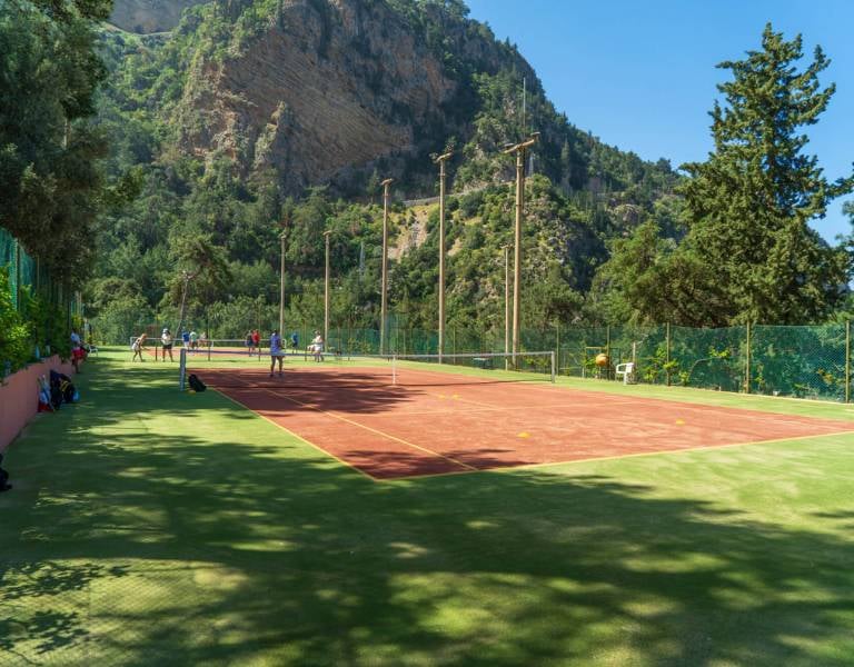 Tennis court at Liberty Lykia Resort with players, set against a backdrop of green mountains and trees, under a clear blue sky.
