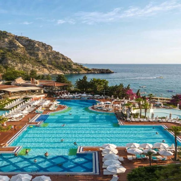Aerial view of Liberty Lykia's large swimming pool surrounded by sun loungers and umbrellas, with the Aegean Sea and mountains in the background.