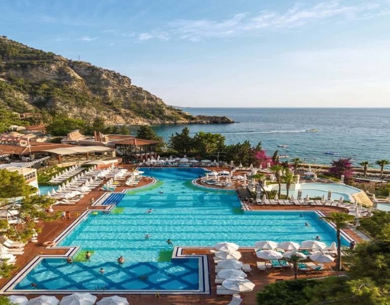 Aerial view of Liberty Lykia's large swimming pool surrounded by sun loungers and umbrellas, with the Aegean Sea and mountains in the background.