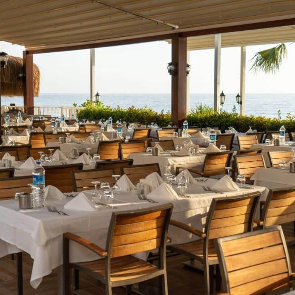 Open-air restaurant at Liberty Lykia Resort with tables set with white tablecloths and wooden chairs, overlooking the sea.