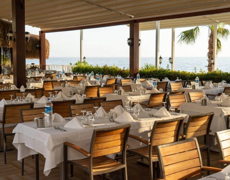 Open-air restaurant at Liberty Lykia Resort with tables set with white tablecloths and wooden chairs, overlooking the sea.