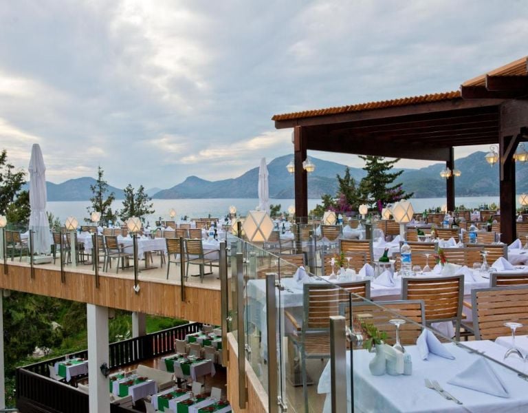 Liberty Lykia rooftop restaurant with sea and mountain views, featuring white tablecloths and wooden seating under a pergola.