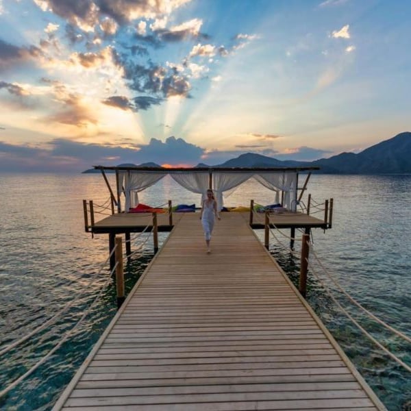 Woman in white atop a wooden pier at sunrise, with sunrays illuminating the sea and mountains at Liberty Lykia.