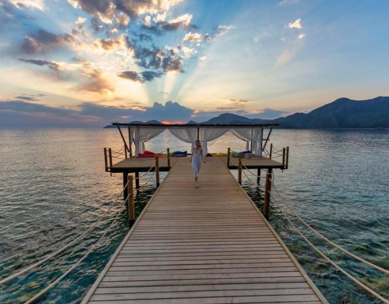 Woman in white atop a wooden pier at sunrise, with sunrays illuminating the sea and mountains at Liberty Lykia.