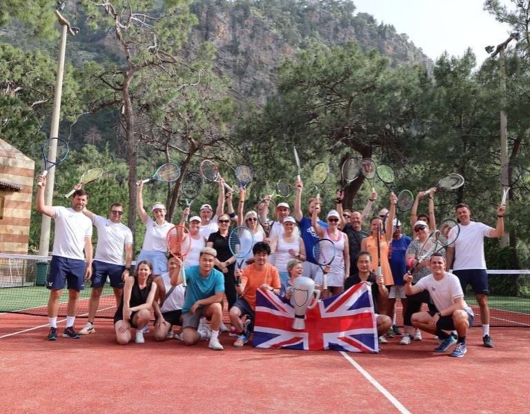 Group of people on a tennis court at Liberty Lykia, holding rackets and a British flag, with trees and mountains in the background.