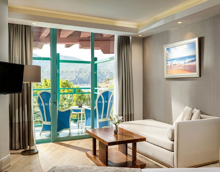 Hotel room at Liberty Lykia Resort with a comfortable lounge area, balcony with blue chairs, and a view of greenery and mountains.