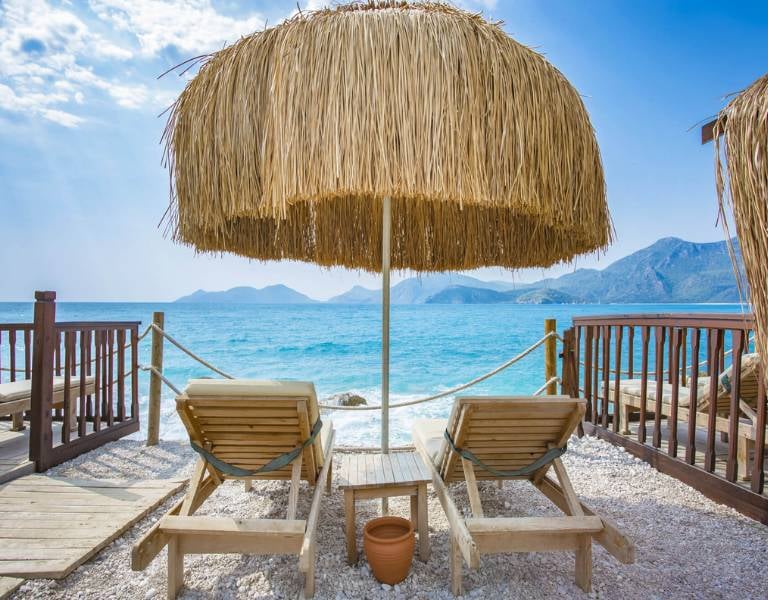 Two loungers on a pebble beach under a straw parasol, facing turquoise sea and mountains at Liberty Sentido Gallery.