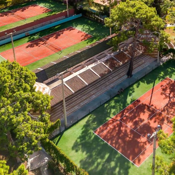 Aerial view of tennis courts among lush trees with players on clay surfaces.