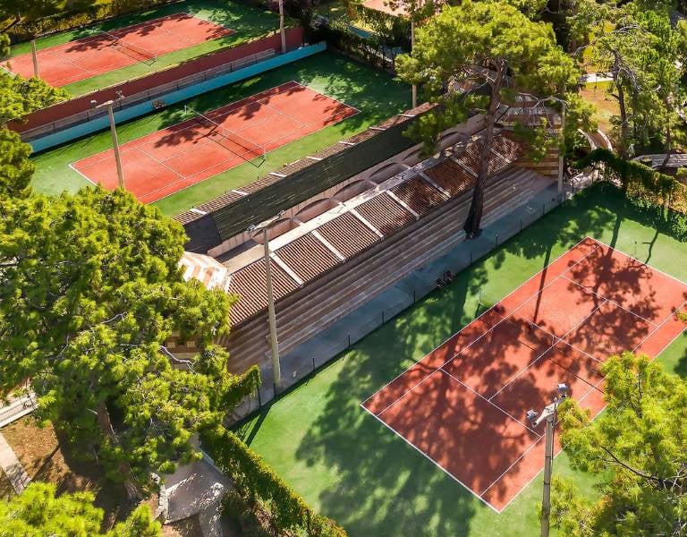 Aerial view of tennis courts among lush trees with players on clay surfaces.
