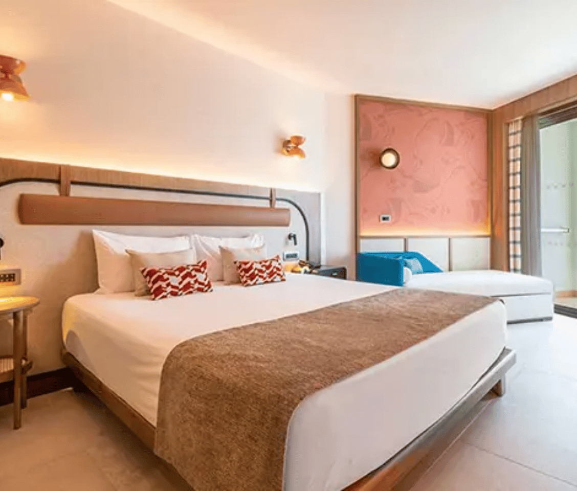 Hotel room at Liberty Village Deluxe with a large double bed, white linens, patterned cushions, and modern decor including a balcony door and wooden accents.