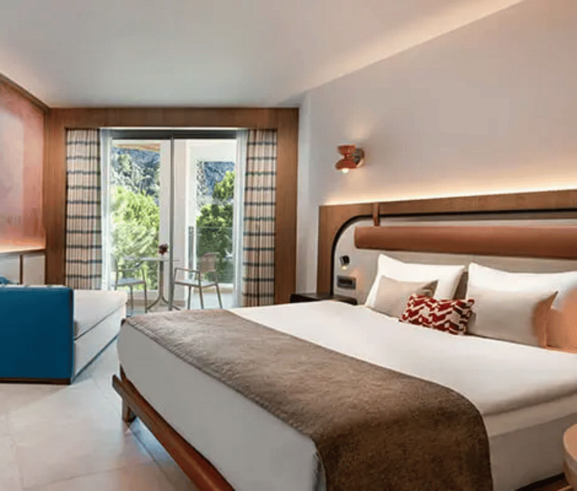 Liberty Village deluxe room with a side sea view, featuring a double bed, blue sofa, and balcony overlooking greenery and sea.