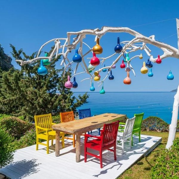 Outdoor seating area at Liberty Village Gallery with a wooden table, colourful chairs, and a tree sculpture decorated with multi-coloured lights, overlooking the sea.