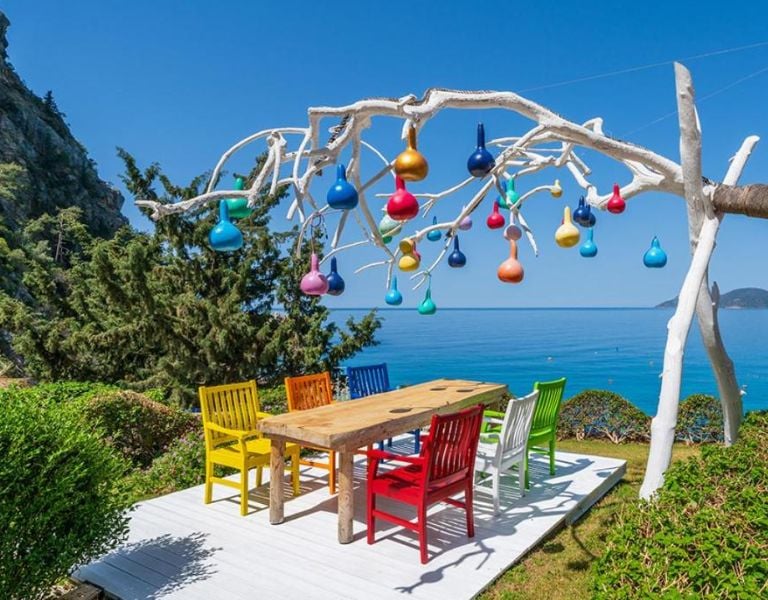 Outdoor seating area at Liberty Village Gallery with a wooden table, colourful chairs, and a tree sculpture decorated with multi-coloured lights, overlooking the sea.