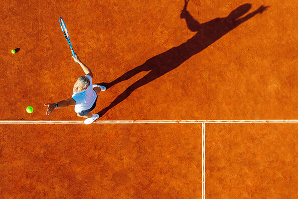 Overhead view of a tennis player serving on a clay court with a racket and two tennis balls, shadow stretched out across the court.