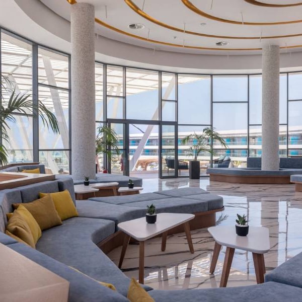 Modern lounge area at Lyttos Gallery with grey sofas, yellow cushions, white tables, and floor-to-ceiling windows with an outdoor view.