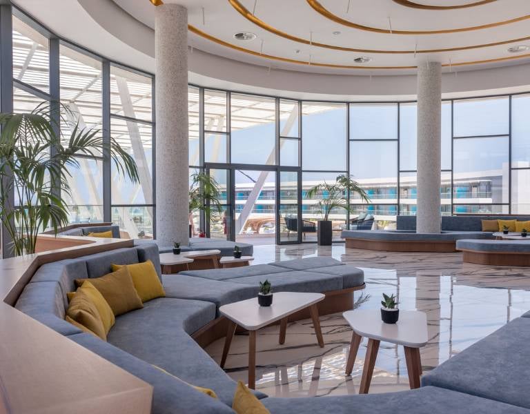 Modern lounge area at Lyttos Gallery with grey sofas, yellow cushions, white tables, and floor-to-ceiling windows with an outdoor view.