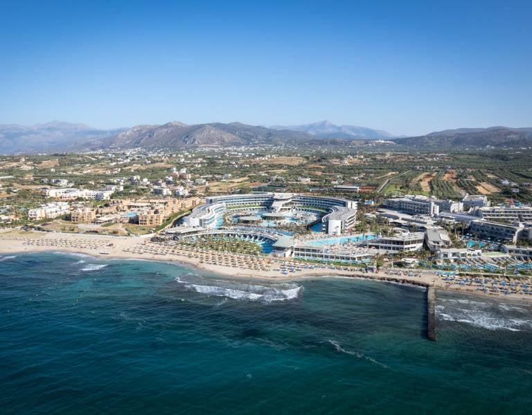 Aerial view of Lyttos Beach Resort in Crete, Greece, showcasing pools, beach, and mountainous backdrop.