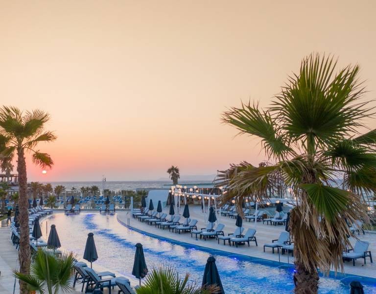 Aerial view of Lyttos Beach hotel pool area at sunset, with an infinity pool, sun loungers, and palm trees.