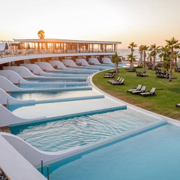 Lyttos Gallery resort pool area at sunset with infinity pools, sun loungers, palm trees, and ocean view.